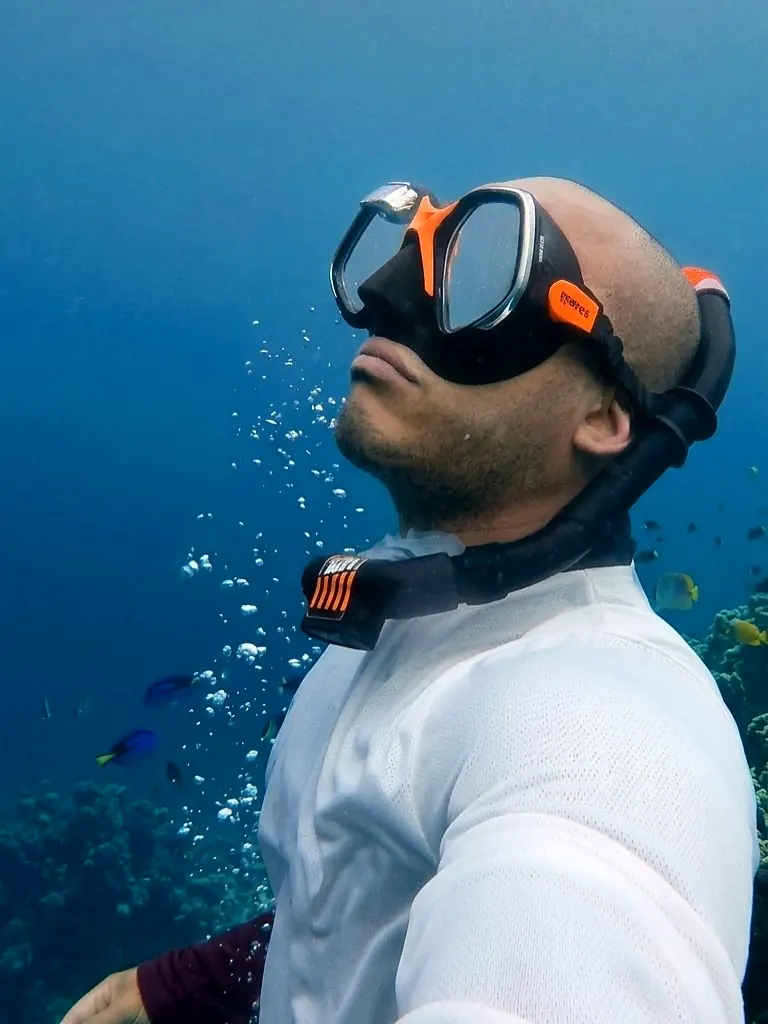Apneísta con máscara naranja mira hacia la superficie rodeado de burbujas y peces